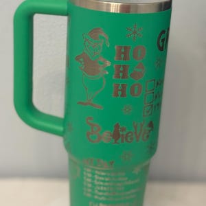 May include: Green insulated tumbler with a handle, featuring a gold-colored Grinch character, snowflakes, and the words "Ho Ho Ho" and "Believe." The tumbler also has a schedule printed on it.