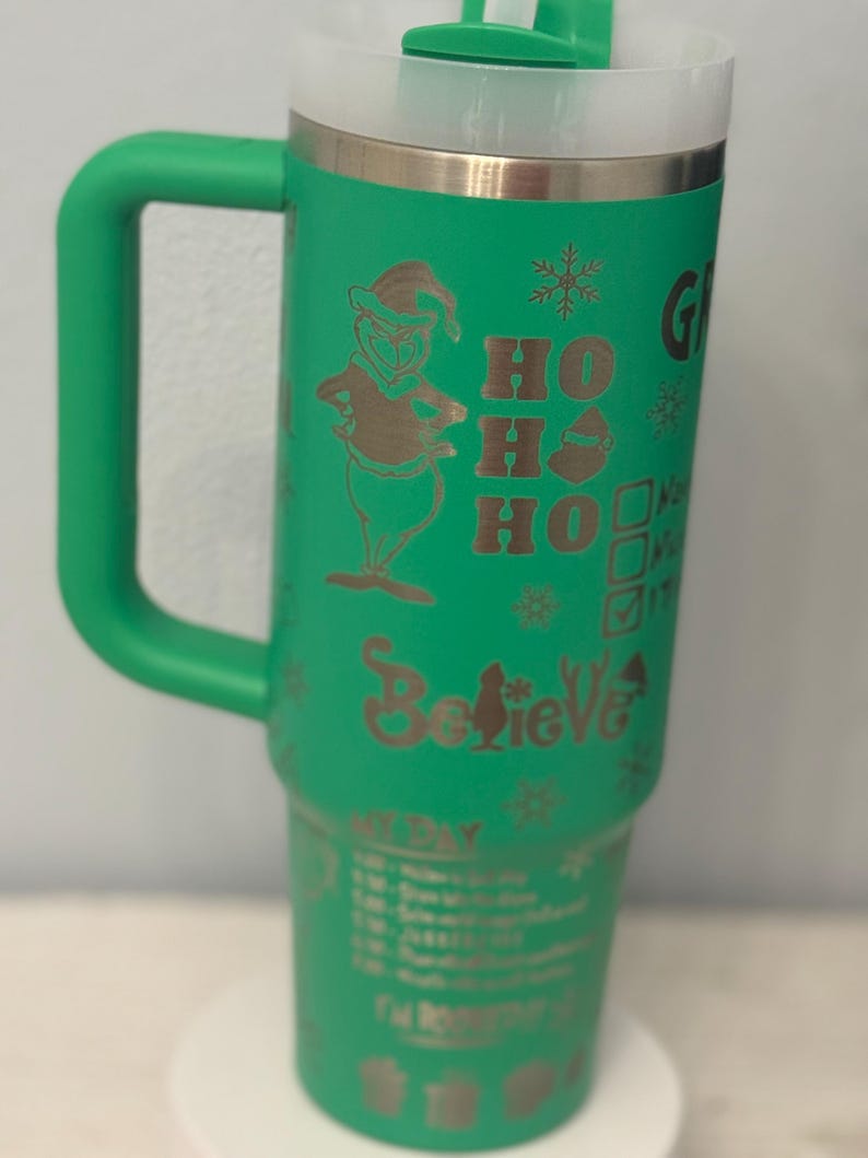 May include: Green insulated tumbler with a handle, featuring a gold-colored Grinch character, snowflakes, and the words "Ho Ho Ho" and "Believe." The tumbler also has a schedule printed on it.