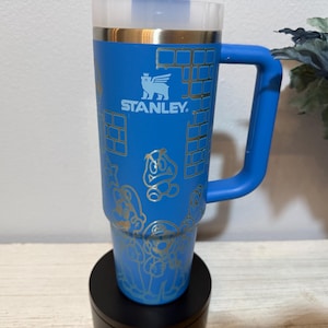 Peut inclure: Gobelet Stanley bleu avec une poign&eacute;e, orn&eacute; de graphiques dor&eacute;s de personnages de jeux vid&eacute;o et de motifs de briques. Le gobelet a un couvercle transparent et un bord dor&eacute;. Le logo Stanley est visible. Le gobelet est sur une base noire.