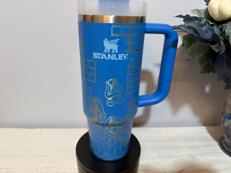 Peut inclure: Gobelet Stanley bleu avec une poign&eacute;e, orn&eacute; de graphiques dor&eacute;s de personnages de jeux vid&eacute;o et de motifs de briques. Le gobelet a un couvercle transparent et un bord dor&eacute;. Le logo Stanley est visible. Le gobelet est sur une base noire.