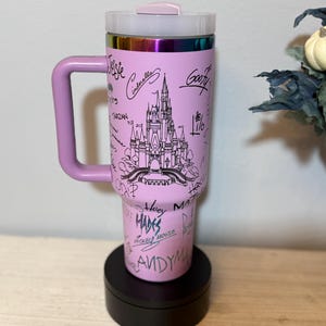 May include: A pink travel mug with a handle, featuring a black line drawing of a castle and character names. The mug has a clear lid and a rainbow-coloured rim. The handle and lid are a matching pink colour.