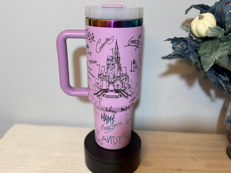 May include: A pink travel mug with a handle, featuring a black line drawing of a castle and character names. The mug has a clear lid and a rainbow-coloured rim. The handle and lid are a matching pink colour.