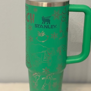 May include: Green insulated travel mug with a handle, featuring a design of the Grinch and snowflakes. The mug has a white lid and the Stanley logo. The mug is approximately 9 inches tall.