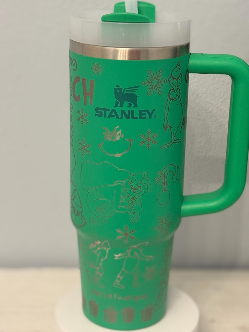 May include: Green insulated travel mug with a handle, featuring a design of the Grinch and snowflakes. The mug has a white lid and the Stanley logo. The mug is approximately 9 inches tall.