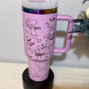May include: A pink travel mug with a handle, featuring signatures of Disney characters like Minnie Mouse and Daisy Duck. The mug has a rainbow-coloured rim and a black base. The mug is designed to keep beverages hot or cold.