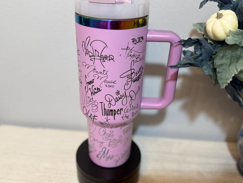 May include: A pink travel mug with a handle, featuring signatures of Disney characters like Minnie Mouse and Daisy Duck. The mug has a rainbow-coloured rim and a black base. The mug is designed to keep beverages hot or cold.
