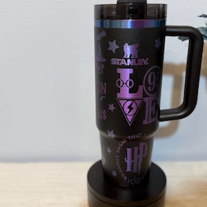 Puede incluir: Taza de viaje Stanley negra con asa, con gráficos y texto morados relacionados con la serie Harry Potter. El diseño incluye estrellas, el logo de Hogwarts y las palabras "LOVE" y "HP". La taza está sobre una base negra.
