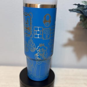 Peut inclure: Un gobelet bleu orn&eacute; d'illustrations dor&eacute;es de personnages et d'&eacute;l&eacute;ments de Super Mario, dont un champignon, des blocs point d'interrogation et la Princesse Peach. Le gobelet est pos&eacute; sur une base noire. Le design rappelle le jeu vid&eacute;o populaire.