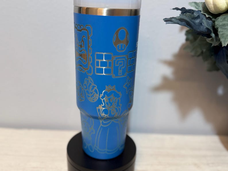 Peut inclure: Un gobelet bleu orn&eacute; d'illustrations dor&eacute;es de personnages et d'&eacute;l&eacute;ments de Super Mario, dont un champignon, des blocs point d'interrogation et la Princesse Peach. Le gobelet est pos&eacute; sur une base noire. Le design rappelle le jeu vid&eacute;o populaire.
