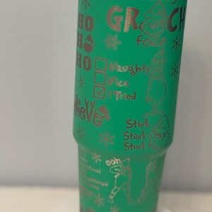May include: A green tumbler with a stainless steel rim and base. The tumbler is decorated with etched images and text, including the words "Resting Grinch Face," snowflakes, and the Grinch character. The tumbler also has the words "Naughty, Nice, I Tried."