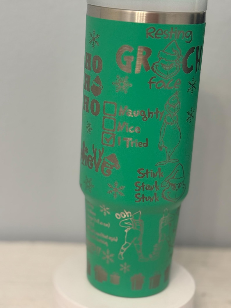 May include: A green tumbler with a stainless steel rim and base. The tumbler is decorated with etched images and text, including the words "Resting Grinch Face," snowflakes, and the Grinch character. The tumbler also has the words "Naughty, Nice, I Tried."
