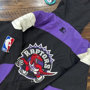 May include: Black, purple, and white Toronto Raptors Starter jacket. The jacket features the Raptors logo on the back, along with the NBA logo on the front. The jacket has a hood and a Starter logo.