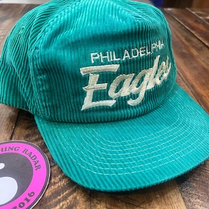 May include: A teal corduroy baseball cap with the text "PHILADELPHIA Eagles" embroidered in white. The cap has a curved brim and a structured crown. A pink and white sticker with the text "VINTAGE CLOTHING RADAR 2016" is in the foreground.