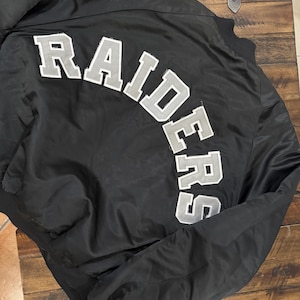 Puede incluir: Chaqueta bomber de satén negro con la palabra "RAIDERS" arqueada en la espalda en letras blancas y grises. La chaqueta tiene cuello, puños y dobladillo de canalé. La chaqueta está sobre una superficie de madera.