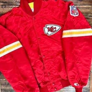Puede incluir: Chaqueta roja de satén con cierre de botones a presión, con el logotipo de los Kansas City Chiefs en el pecho y un parche de la NFL en la manga. La chaqueta tiene rayas blancas y amarillas en las mangas y cuello y puños rojos.