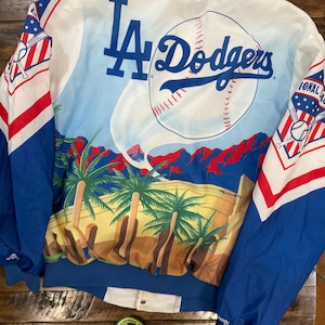 Puede incluir: Chaqueta de béisbol blanca y azul de los Dodgers de Los Ángeles con una colorida escena desértica en la espalda. La chaqueta presenta el logotipo del equipo y un gráfico de béisbol. Las mangas tienen detalles en rojo, blanco y azul.