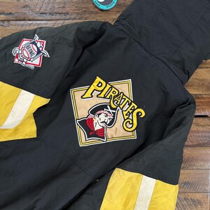 Puede incluir: Chaqueta de béisbol negra y amarilla con capucha. La chaqueta presenta un logotipo de los Piratas en la espalda y un parche de la Liga Nacional en la manga. La chaqueta tiene rayas blancas en las mangas.
