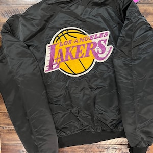 Puede incluir: Chaqueta bomber de satén negro con el logotipo de Los Angeles Lakers en amarillo y morado. La chaqueta tiene cuello, puños y dobladillo de canalé negro. Un logotipo de Starter es visible en la manga izquierda.