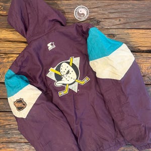 Op de afbeelding: Een paarse Starter-jas met capuchon, met turquoise en witte accenten op de mouwen. De jas toont het Anaheim Ducks-logo op de rug, samen met het NHL-logo op de mouw. De jas ligt op een houten oppervlak.