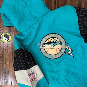 May include: Teal and black vintage Florida Marlins hooded jacket. The jacket features the team logo on the back and a black and white striped accent on the sleeve. The jacket is made of a lightweight material and has a hood.