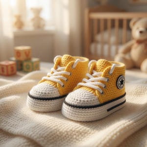 May include: A pair of handmade yellow and white crochet baby sneakers with white laces and a black sole. The sneakers have a small black and white patch with a star. The shoes are on a cream-colored blanket.
