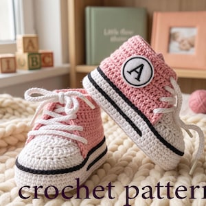 Può includere: Un paio di scarpine da neonato all'uncinetto rosa e bianche con lacci bianchi e una striscia nera. Una scarpa ha una toppa con la lettera "A". Il testo "crochet pattern" è in basso nell'immagine. Le scarpe sono su una coperta color crema.
