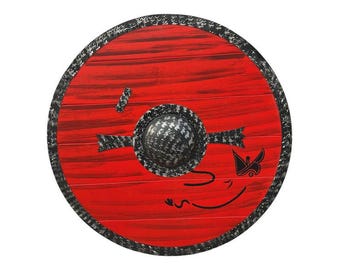 May include: A round, red wooden shield with a black and silver patterned rim and central boss. The shield features a black design of a stylized face and other decorative elements. The shield is a decorative item.