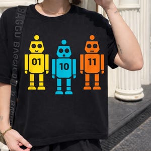 May include: Black t-shirt featuring three colorful robot illustrations. The robots are yellow, blue, and orange, each with a unique number: 01, 10, and 11. The design is simple and graphic, perfect for a casual look.