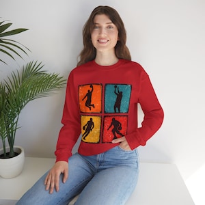 May include: A red sweatshirt with a basketball design. The design features four squares, each with a silhouette of a basketball player in action, set against a colourful, distressed background. The sweatshirt is a solid red colour.