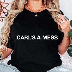 Könnte beinhalten: Schwarzes T-Shirt mit Rundhalsausschnitt und dem weißen Text "CARL'S A MESS". Das T-Shirt wird von einer Person mit langen blonden Haaren getragen, die eine Halskette und Ringe trägt. Die Person sitzt.