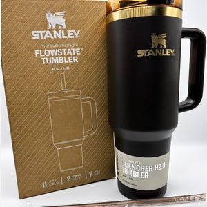 May include: Black Stanley Flowstate tumbler with a gold rim and handle, beside its cardboard box. The box displays the Stanley logo and product details, including "48 OZ | 1.18L". A reusable straw is also visible.