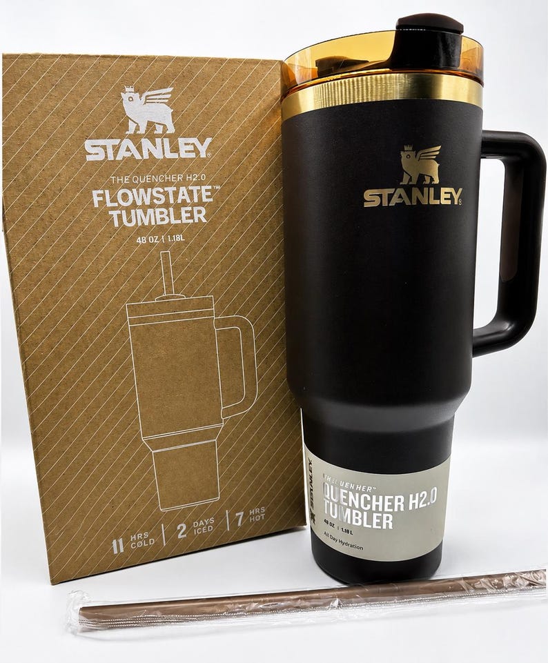 May include: Black Stanley Flowstate tumbler with a gold rim and handle, beside its cardboard box. The box displays the Stanley logo and product details, including "48 OZ | 1.18L". A reusable straw is also visible.