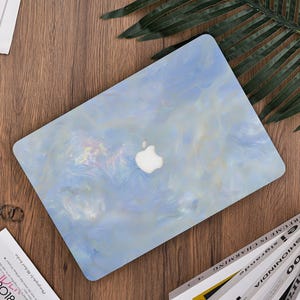 Puede incluir: Funda para portátil veteada en azul claro y blanco con el logotipo de Apple. La funda está sobre una superficie de madera con revistas y una hoja de palma. La funda tiene un acabado nacarado.