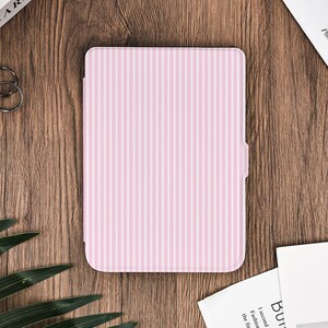 Pode incluir: Uma capa para tablet com listras rosa e brancas. A capa é retangular com um padrão de listras verticais. A capa está fechada e parece ser feita de um material liso. O fundo é uma superfície de madeira com livros e outros itens.
