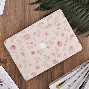 May include: A laptop case adorned with a floral design of pink roses and foliage on a pale pink backdrop. The case rests on a wooden surface, accompanied by a palm leaf and magazines.