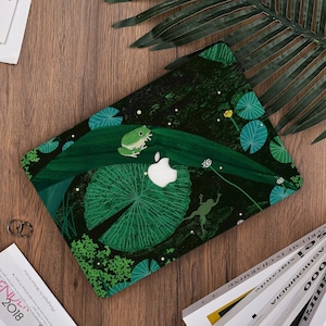 May include: Laptop case with a green frog and lily pad design. The case features a white Apple logo and a dark green background with a nature theme. The design includes a frog on a leaf and other aquatic plants.