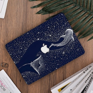 Könnte beinhalten: Eine marineblaue Laptop-Hülle mit einer weißen Illustration eines Mantarochens und eines Walhais, die durch einen sternenübersäten Nachthimmel schwimmen. Das Apple-Logo ist sichtbar. Die Hülle liegt auf einer Holzoberfläche.