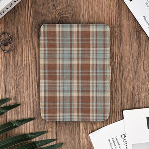 Peut inclure: Un étui rectangulaire avec un motif à carreaux marron, bleu et beige. L'étui est posé sur une surface en bois. Il est probablement destiné à un appareil électronique, comme une tablette ou une liseuse.