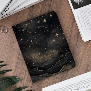 Könnte beinhalten: Schwarzes Notizbuch mit einem Himmelsdesign mit goldenen Sternen und einem dunklen, strukturierten Hintergrund. Das Notizbuch hat eine abgerundete Kante und liegt auf einer Holzoberfläche. Weitere Artikel sind Ringe und Drucksachen.