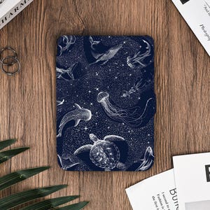 Könnte beinhalten: Eine dunkelblaue Tablet-Hülle mit einer weißen Illustration verschiedener Meeresbewohner, darunter Wale, Quallen und Schildkröten, vor einem Sternenhimmel. Die Hülle ist rechteckig und hat einen Magnetverschluss.