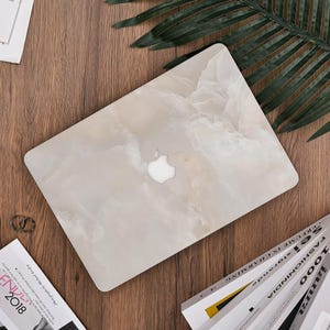 Könnte beinhalten: Ein Laptop mit einem hellbeigen marmorähnlichen Design. Das Apple-Logo ist auf dem Cover sichtbar. Der Laptop liegt auf einer Holzoberfläche, mit einem grünen Blatt und Zeitschriften in der Nähe.