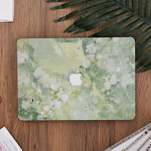 以下が含まれることがあります： 緑と白の花柄のノートパソコンケース。ケースは、中央に白いAppleのロゴが付いた水彩画風のパターンが特徴です。デザインは、緑、白、青の色合いを取り入れています。