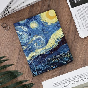 Può includere: Un taccuino con una copertina raffigurante "La notte stellata" di Vincent van Gogh. Il design mostra i vorticosi blu, gialli e bianchi del dipinto. Il taccuino rettangolare è appoggiato su una superficie di legno.