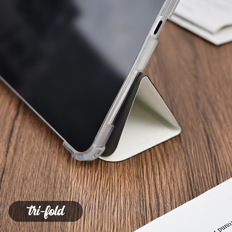 Pu&ograve; includere: Una custodia trasparente per tablet con supporto tri-fold. Il tablet &egrave; appoggiato su una superficie di legno. La custodia &egrave; trasparente e il supporto &egrave; bianco e nero. Il testo "tri-fold" &egrave; in un ovale marrone.