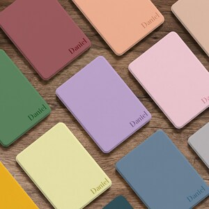 May include: A collection of rectangular, solid-colored passport holders in shades of maroon, peach, beige, green, lavender, light pink, yellow, blue, and gray. Each holder features the name Daniel embossed in a dark, sans-serif font on the front.
