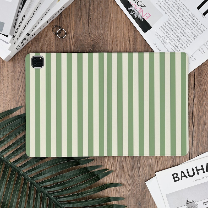 Pu&ograve; includere: Una custodia rettangolare per tablet con un motivo a righe verticali in verde chiaro e bianco sporco. La custodia &egrave; appoggiata su una superficie di legno. Un obiettivo della fotocamera &egrave; visibile nell'angolo in alto a sinistra.