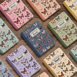 Può includere: Una collezione di quaderni rettangolari con varie copertine color pastello decorate con motivi di farfalle e fiori. Ogni quaderno presenta un'etichetta bianca centrale con il testo "COMPOSITION BOOK" e il nome "Daniel" stampato in nero.
