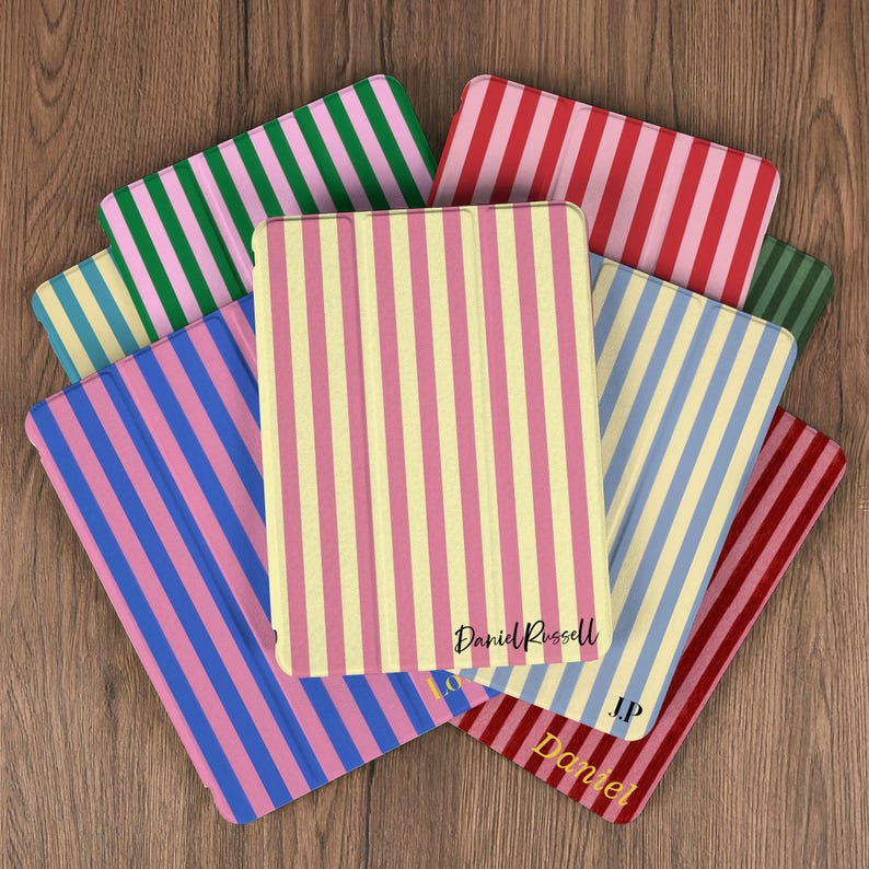 Pu&ograve; includere: Una collezione di custodie per tablet colorate con motivi a righe verticali in vari colori, tra cui rosa, giallo, blu, verde e rosso. Le custodie hanno il nome "Daniel Russell" stampato.