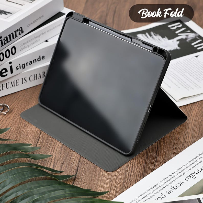 Pu&ograve; includere: Una custodia nera per tablet con la scritta "Book Fold" in un ovale marrone. La custodia &egrave; aperta, rivelando uno schermo nero. La custodia &egrave; su una superficie di legno con una pila di libri e un giornale sullo sfondo.