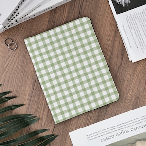 May include: A green and white gingham pattern notebook or tablet cover. The cover is rectangular with rounded corners and a classic checkered design. The cover is lying on a wooden surface, with other objects in the background.